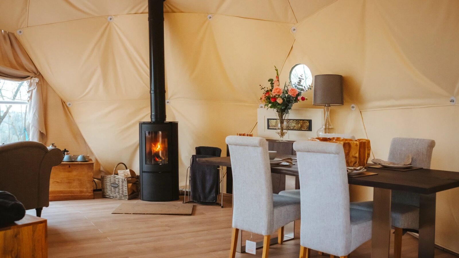 Experience the charm of Woodland Escapes in a cozy glamping dome, featuring a wood-burning stove, dining table, chairs, an armchair, and soft lighting.