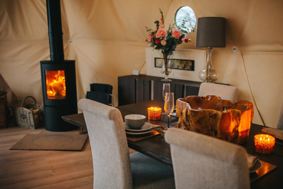 A cozy dining area awaits, featuring a wooden table and chairs bathed in candlelight. Nearby, a wood stove warms the space, while a bouquet of flowers sits gracefully beside the lamp—a perfect Woodland Escapes retreat.