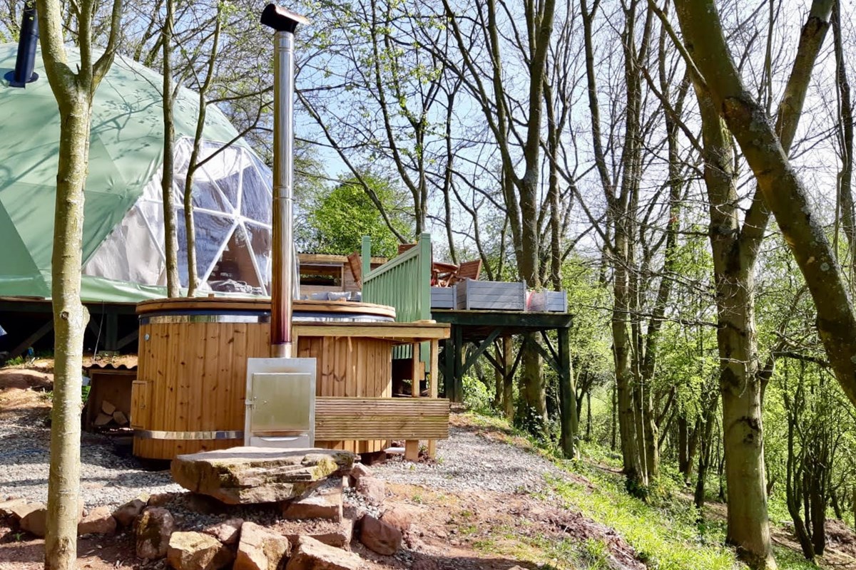 Experience the tranquility of Woodland Escapes with a wooden hot tub and green dome tent nestled amidst trees, complete with a raised deck in a serene forest setting.