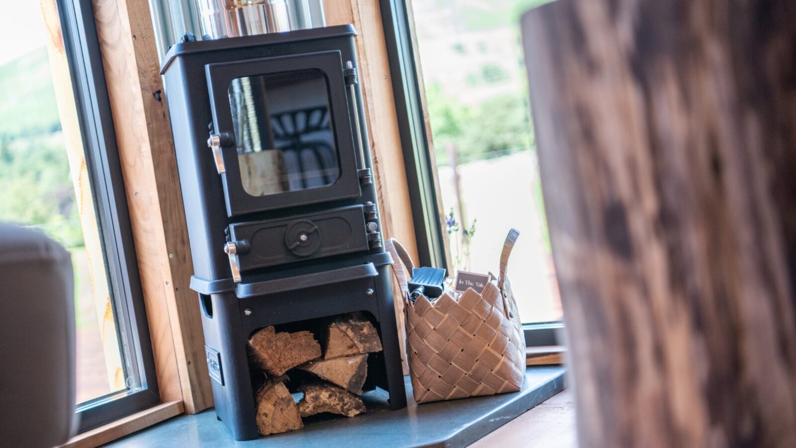 A black wood-burning stove with firewood stacked below sits in a cozy room, overlooking The Vale through large windows, accompanied by a charming wicker basket.