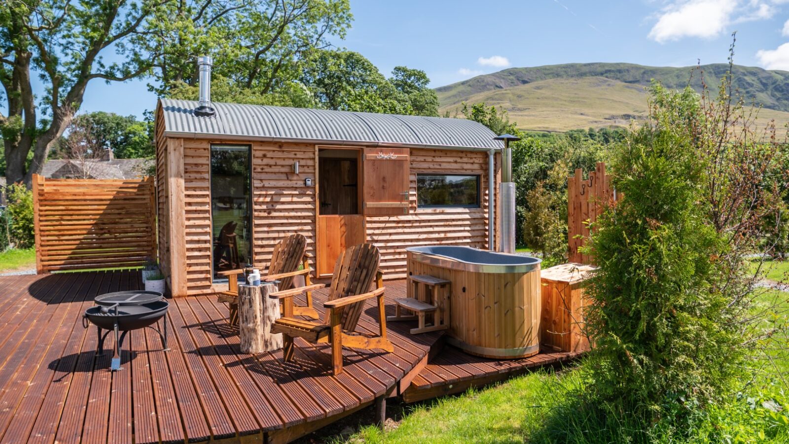 Nestled in The Vale, this charming small wooden cabin features a deck with two chairs, inviting guests to soak in the hot tub or enjoy evenings by the fire pit amid a picturesque grassy area and rolling hills.