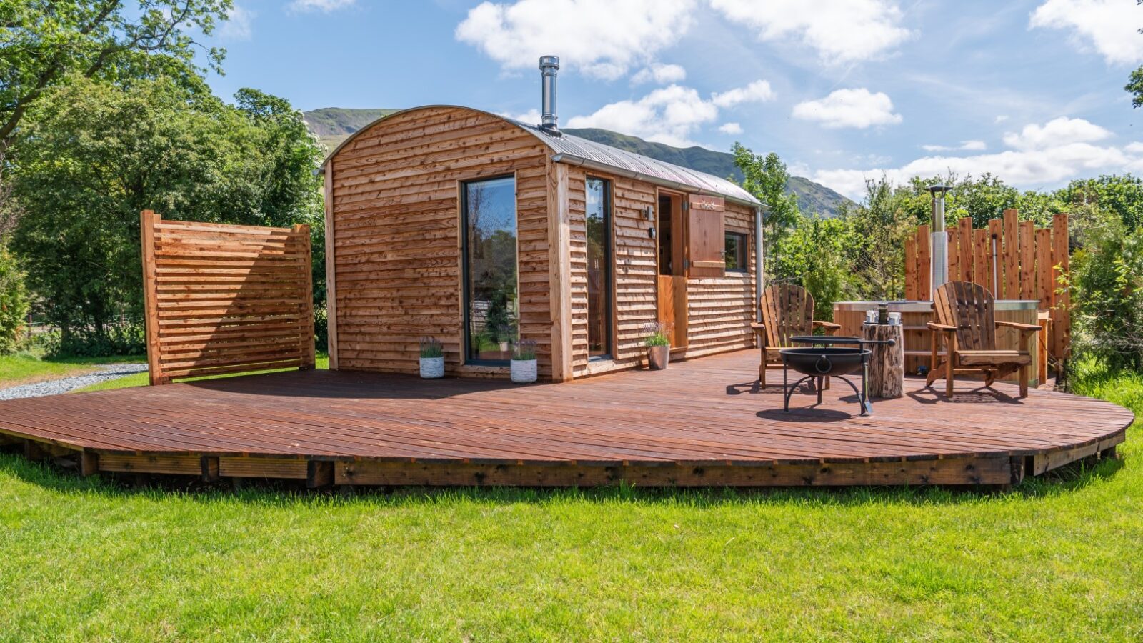 A small wooden cabin with a curved roof sits on a raised deck, nestled in the lush greenery of The Vale under a partly cloudy sky.