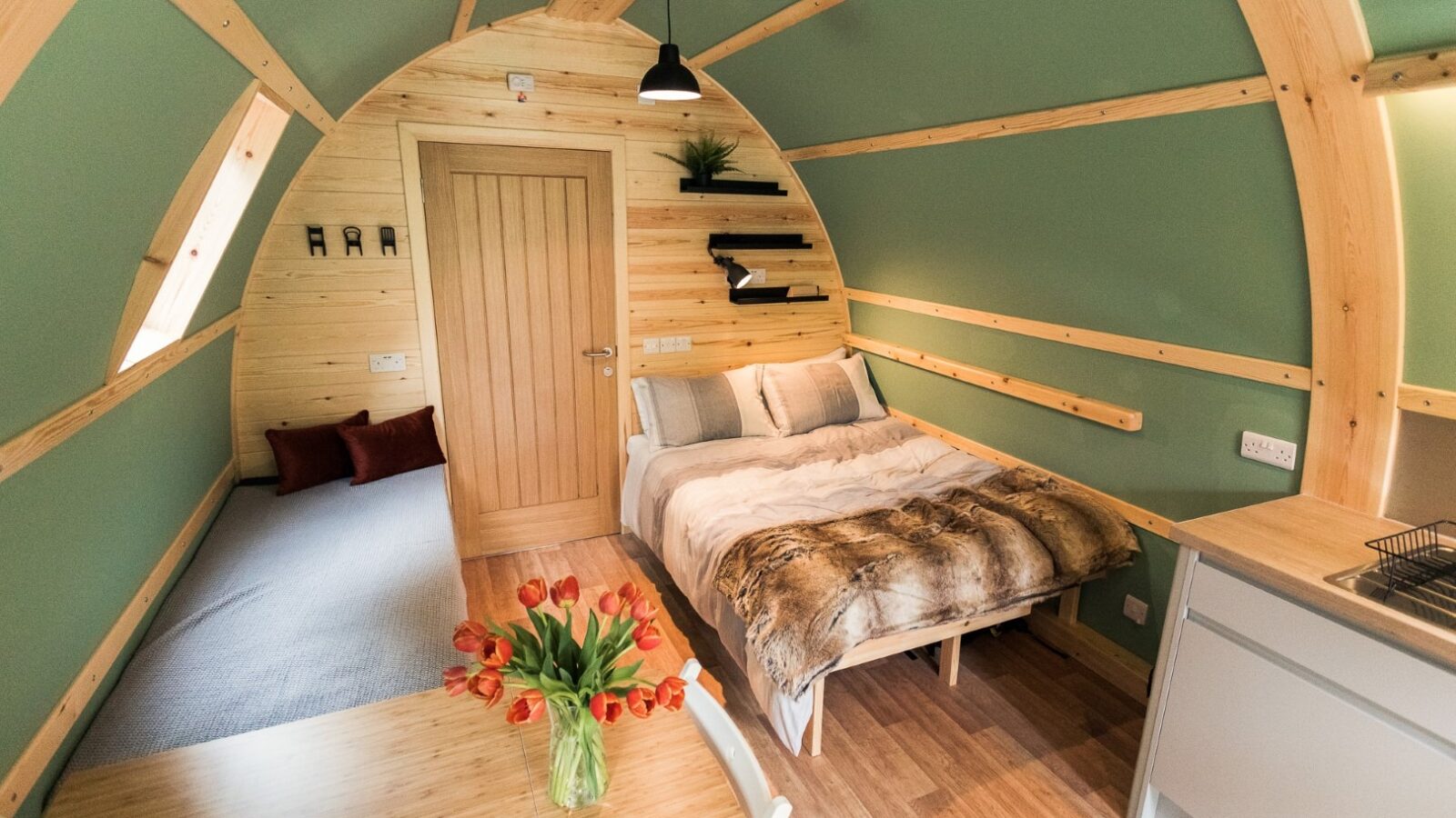 A cozy, small wooden cabin interior reminiscent of Forcett Grange, with a curved ceiling and sage-green walls. It features a bed with fur-lined bedding, a small sofa, a kitchenette with a sink, and a wooden table holding a vase of orange tulips. Shelves are mounted above the bed.