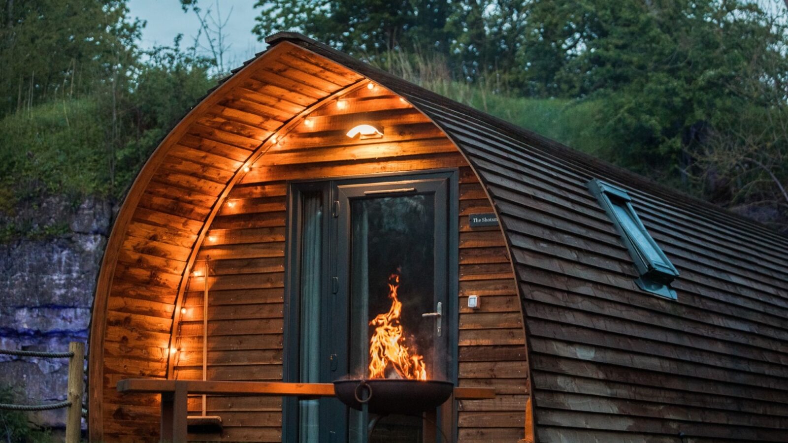 A cozy wooden cabin with warm yellow lights strung along the roofline stands gracefully at Forcett Grange. The cabin, featuring a curved roof, nestles in a lush, green outdoor setting. A fire pit with a bright, crackling flame sits in front of the cabin, next to a wooden picnic table.
