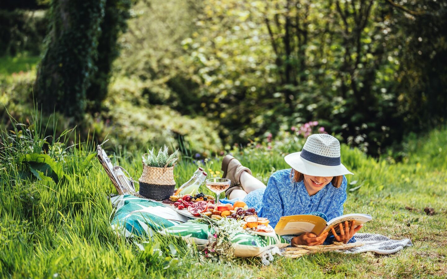 A person in a blue dress and sunhat lounges on a blanket, absorbed in a book. Nearby, a picnic spread of fruits, veggies, bread, and wine rests invitingly. The serene surroundings evoke the charm of Devon's tranquil landscapes, with trees and greenery enveloping the scene.