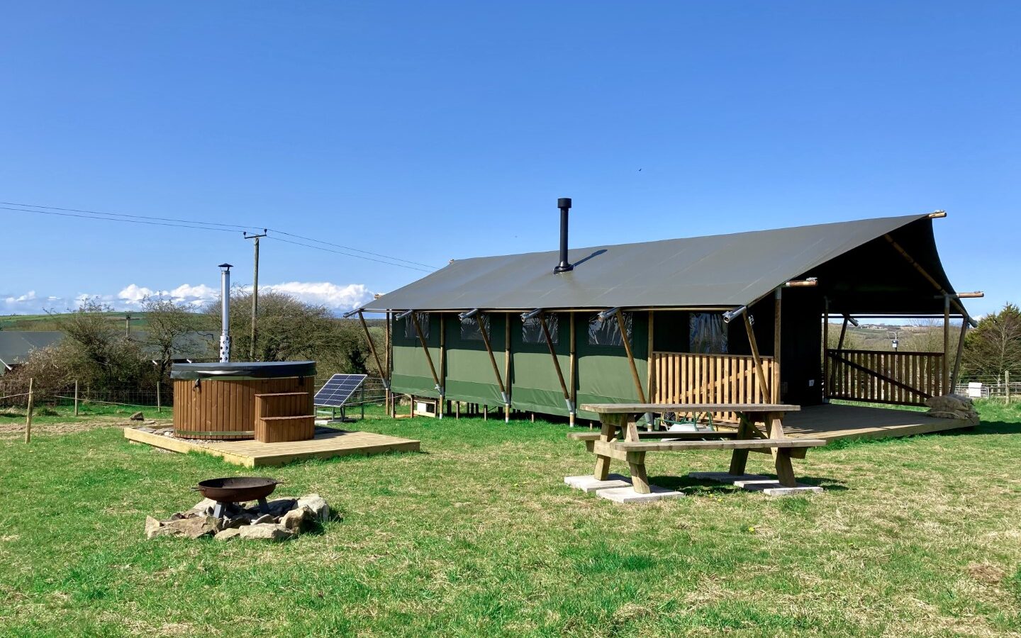 At Sloeberry Farm, a safari-style lodge with a chimney rests on a lush grass field, accompanied by a wooden hot tub, picnic table, and fire pit under the expansive blue sky.