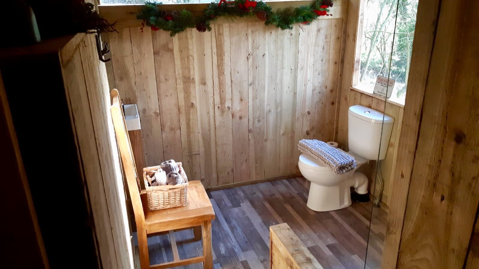 This Woodland Escapes cabin bathroom features a toilet, a chair with a basket, and a window adorned with a pine garland above.