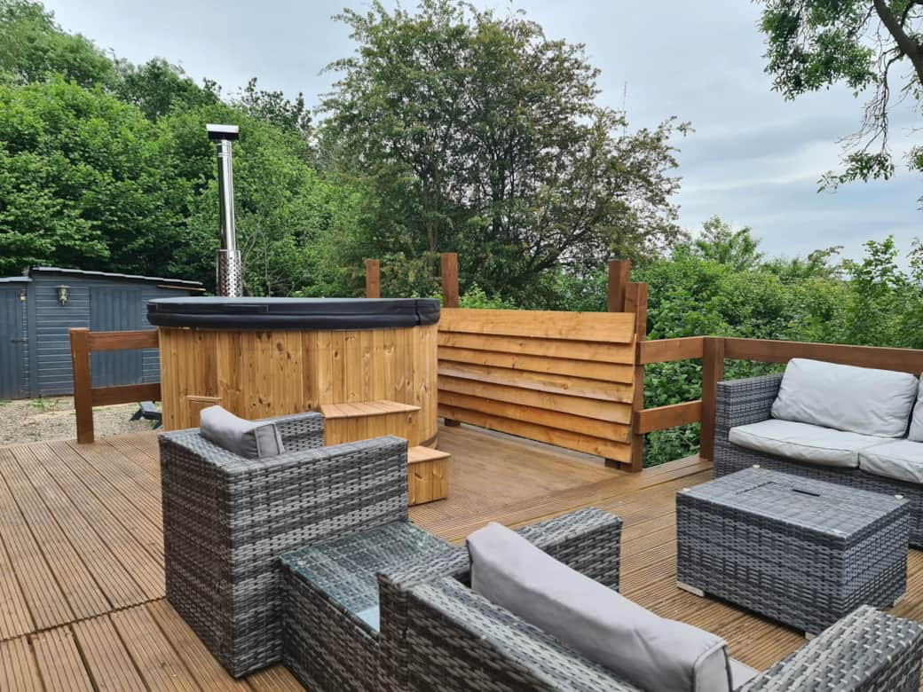 A serene woodland escape featuring a wooden deck with a hot tub, surrounded by wicker furniture and cushions, all nestled amidst trees under a cloudy sky – the perfect lodge-like retreat.