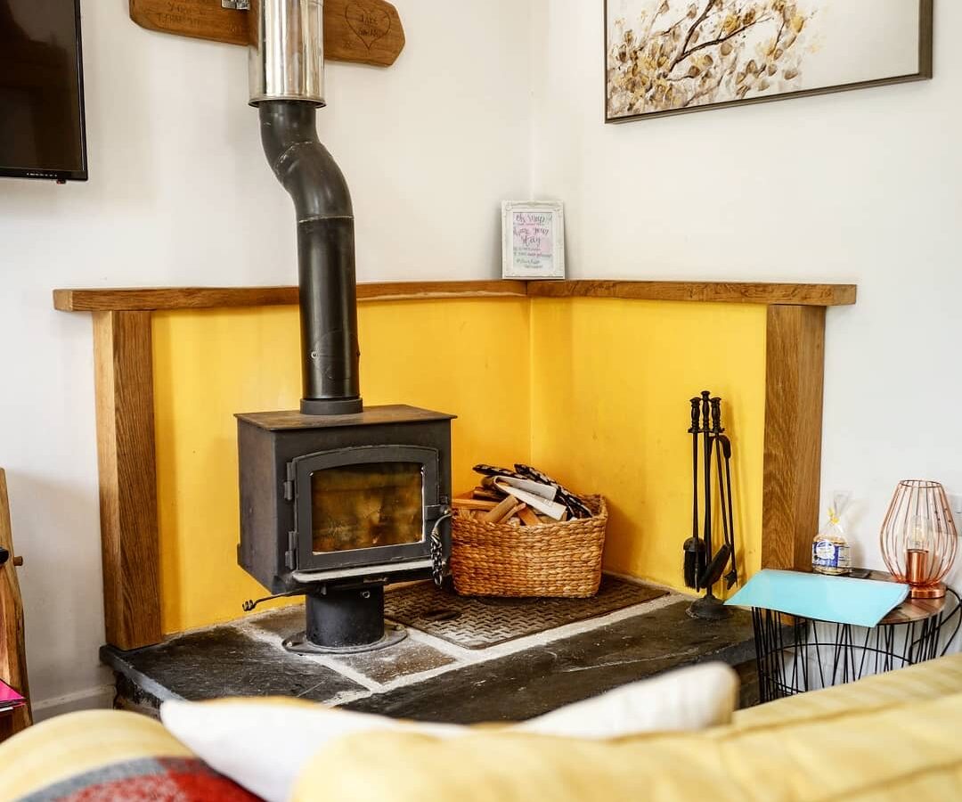 Cozy room at Nant Awen Cottages with a wood-burning stove, wicker basket, and fireplace tools. The yellow accent wall and framed art above add charm to this inviting space.