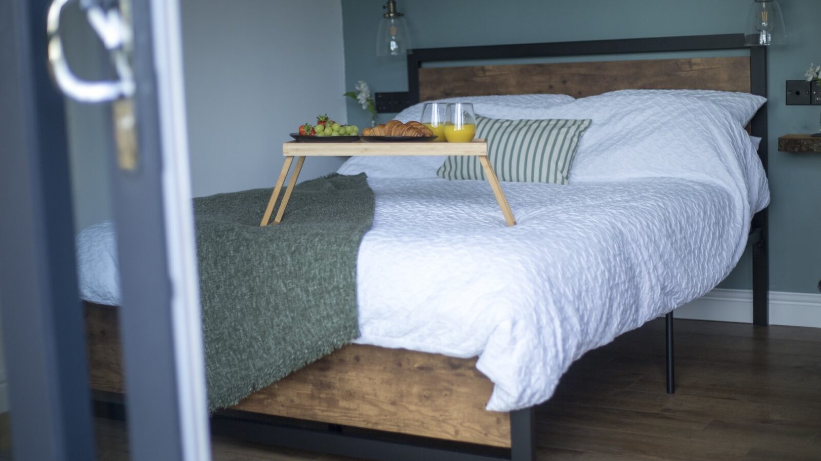 A cozy cabin bedroom features a neatly made bed with a white quilt and green throw. A breakfast tray with food and drinks is placed on the bed, beneath a pair of hanging pendant lights. The décor includes a rustic wood and metal headboard, complementing the Cae Cnwc charm.