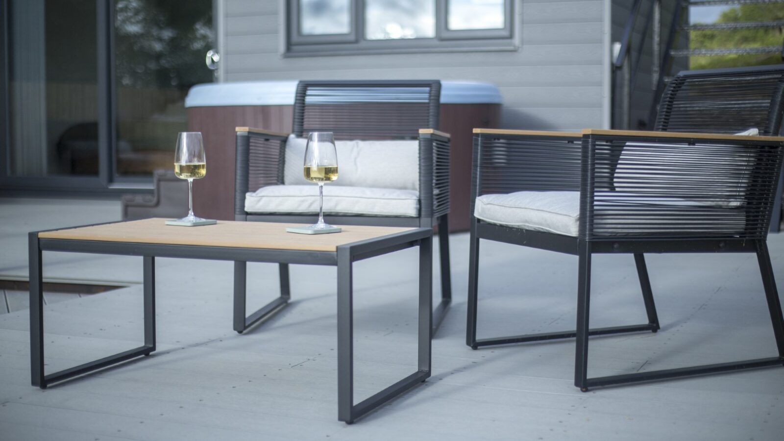 A modern outdoor patio setup at Cae Cnwc Cabin features two black and wood chairs with white cushions, a matching table, and two glasses of white wine. The background includes a hot tub and stairs leading up to the inviting house.