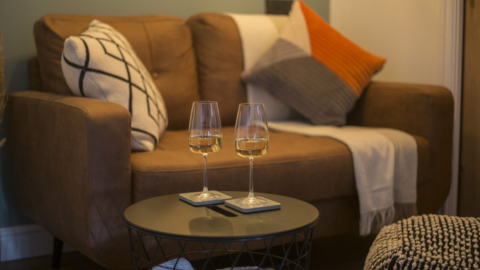 A cozy living room in Cae Cnwc Cabin features a brown sofa adorned with patterned pillows and a throw. Two wine glasses rest on a black wire-frame coffee table. A pouffe and soft blanket complete the inviting atmosphere.