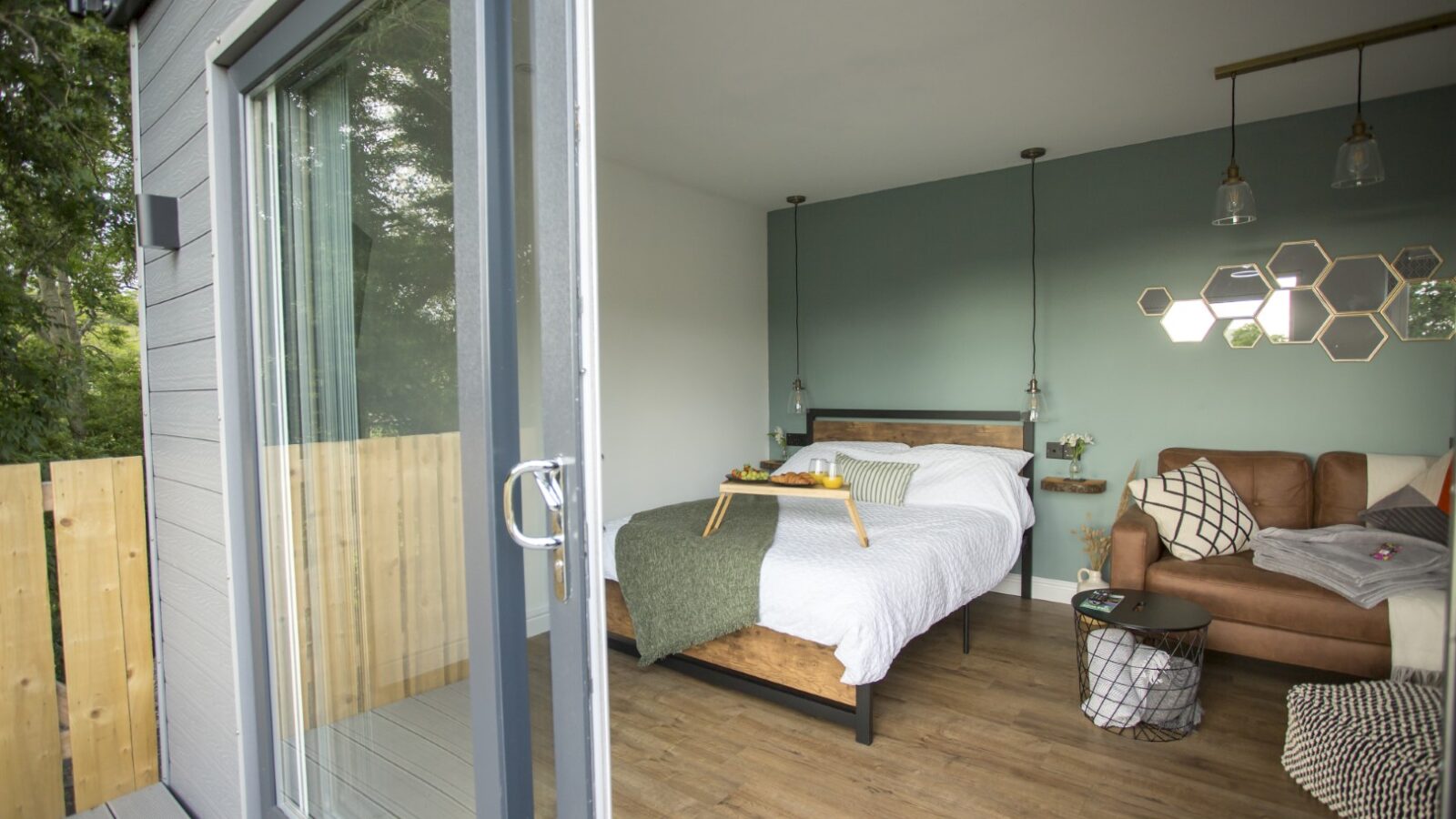 A cozy room in Cae Cnwc Cabin features a bed against a green accent wall and wooden flooring. A tray with breakfast awaits on the bed. Nearby, a brown couch and coffee table enhance the hexagonal wall decor, while the sliding glass door leads to a deck offering a fenced view.