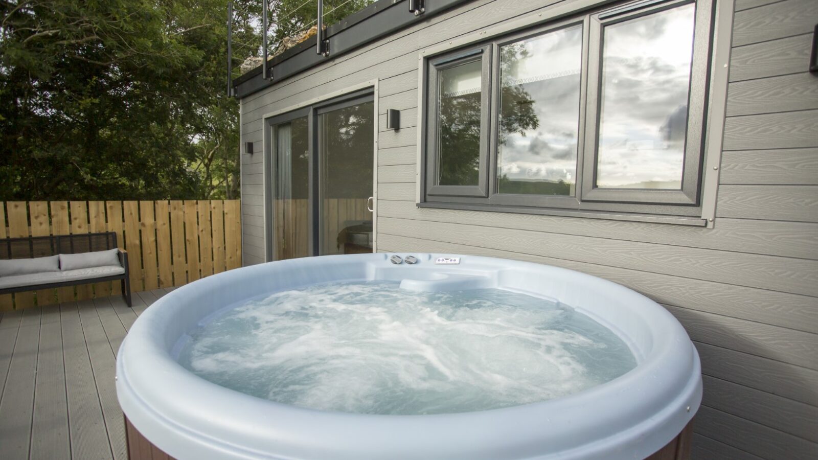 A round hot tub with swirling water sits on a wooden deck in front of the modern gray Cae Cnwc Cabin, featuring large windows. Trees and a wooden fence are visible in the background, under a cloudy sky.