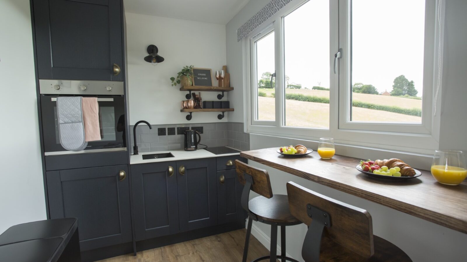 A cozy kitchen in the Cae Cnwc Cabin features dark cabinets, a wall-mounted oven, and a small sink. A wooden breakfast bar with two stools overlooks large windows, offering a view of a grassy field. Plates with croissants and fruit sit beside glasses of orange juice.