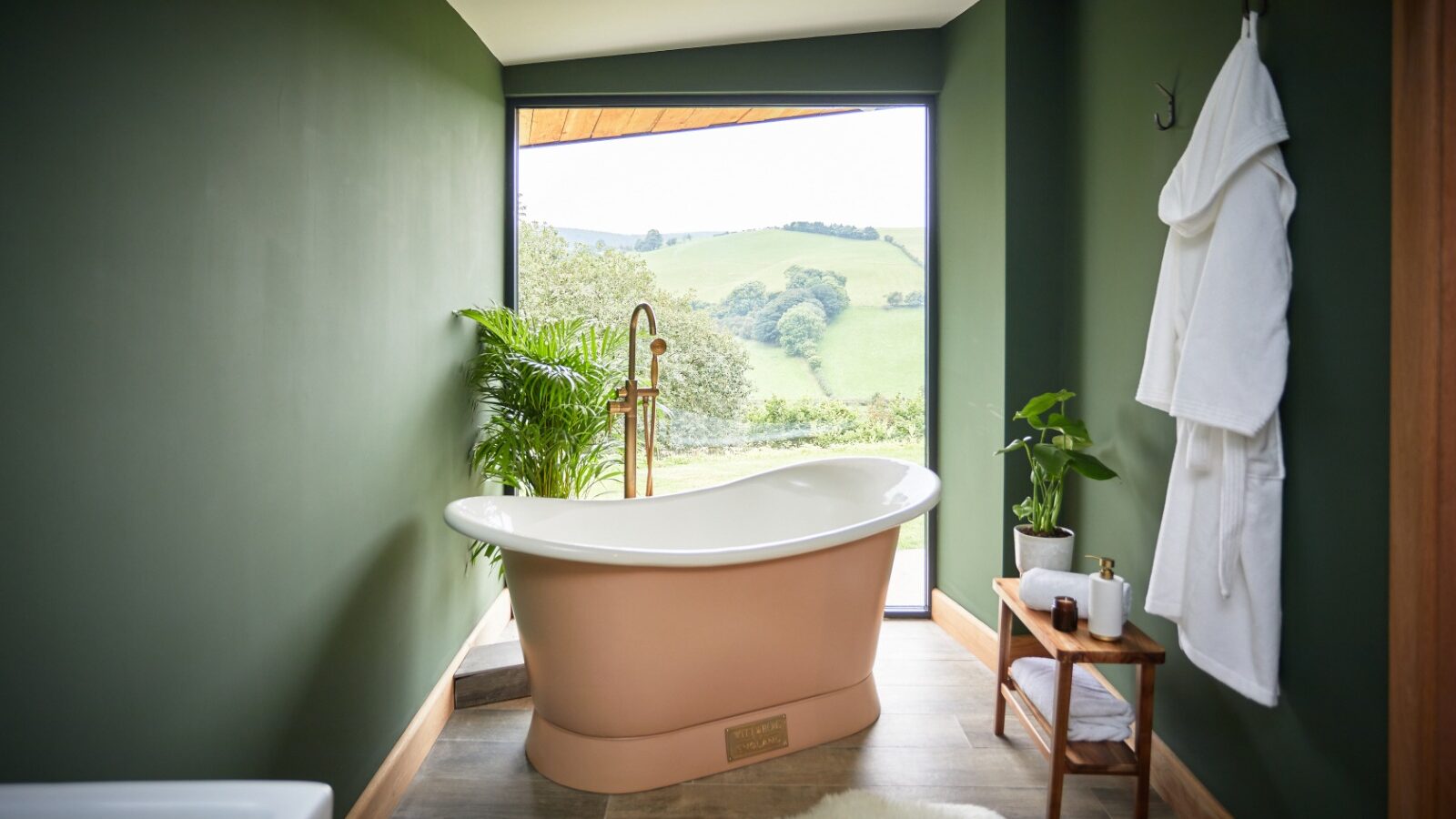 A tranquil bathroom from Burrows features a freestanding tub set against a large window overlooking a lush green landscape. The room has Dolassey green walls, wooden flooring, a fluffy white rug, a potted plant, and bathrobes hanging on the wall.