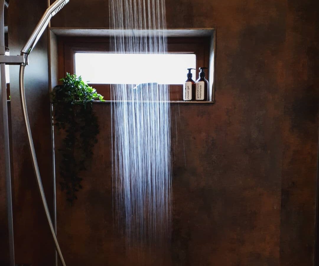 A modern shower with a Burrows rainfall showerhead cascades down. Above, a rectangular window holds two bottles and a small green plant on the ledge. The Dolassey-style shower walls boast a dark, textured finish, enhancing the sleek ambiance.