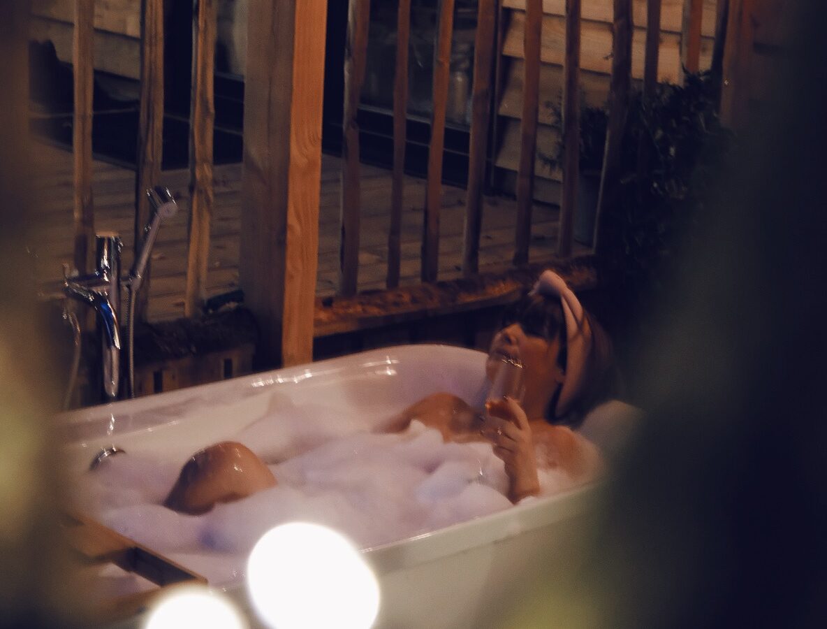 Beneath the branches, a person unwinds in a bubble bath outdoors, enveloped by wooden structures and soft lighting at night.