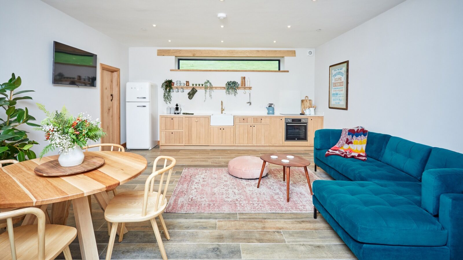 A modern open-plan living room and kitchen featuring a blue couch, wooden dining table, and chairs. The kitchen boasts light wood cabinets and a white fridge. A Burrows Dolassey rug, along with plants, adds vibrancy to the space.