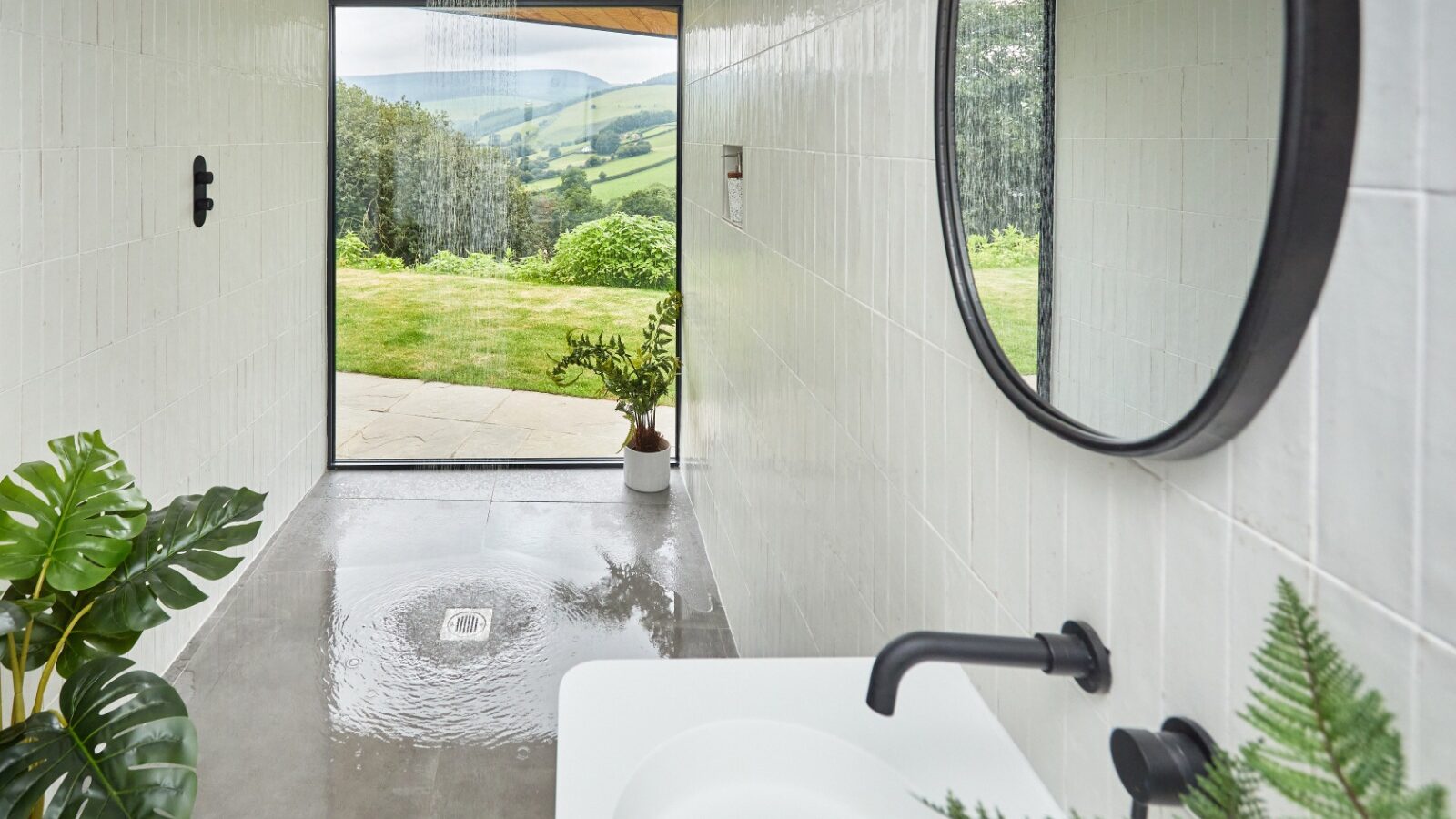 A modern bathroom with white tiled walls, featuring a round mirror and sleek black fixtures. Glass doors open out to a scenic green landscape with rolling hills, reminiscent of the serene beauty of Burrows. A rainy showerhead and lush potted plants enhance the natural tranquility.