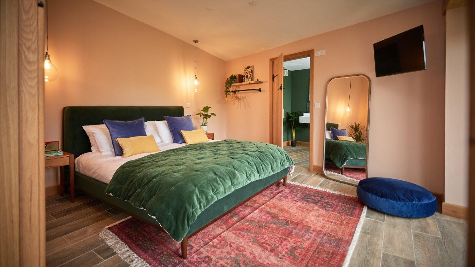 A cozy bedroom featuring a green velvet bed adorned with colorful pillows and a warm-toned rug. A Burrows Dolassey floor mirror leans against the wall, while a blue cushion rests on the wooden floor. Pendant lights hang from the ceiling, casting a gentle glow.