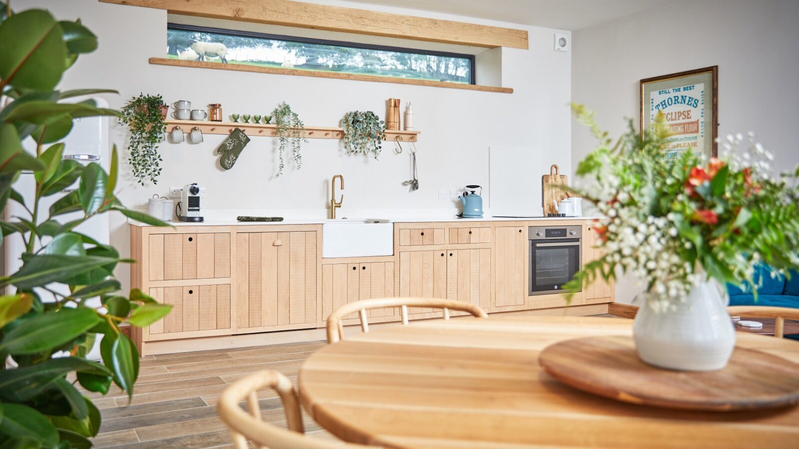 A modern kitchen in The Burrows features light wood cabinets, a farmhouse sink, and floating shelves adorned with plants and kitchenware. A round wooden table with a floral centerpiece graces the foreground, while a large window in this Dolassey-designed space provides ample natural light.