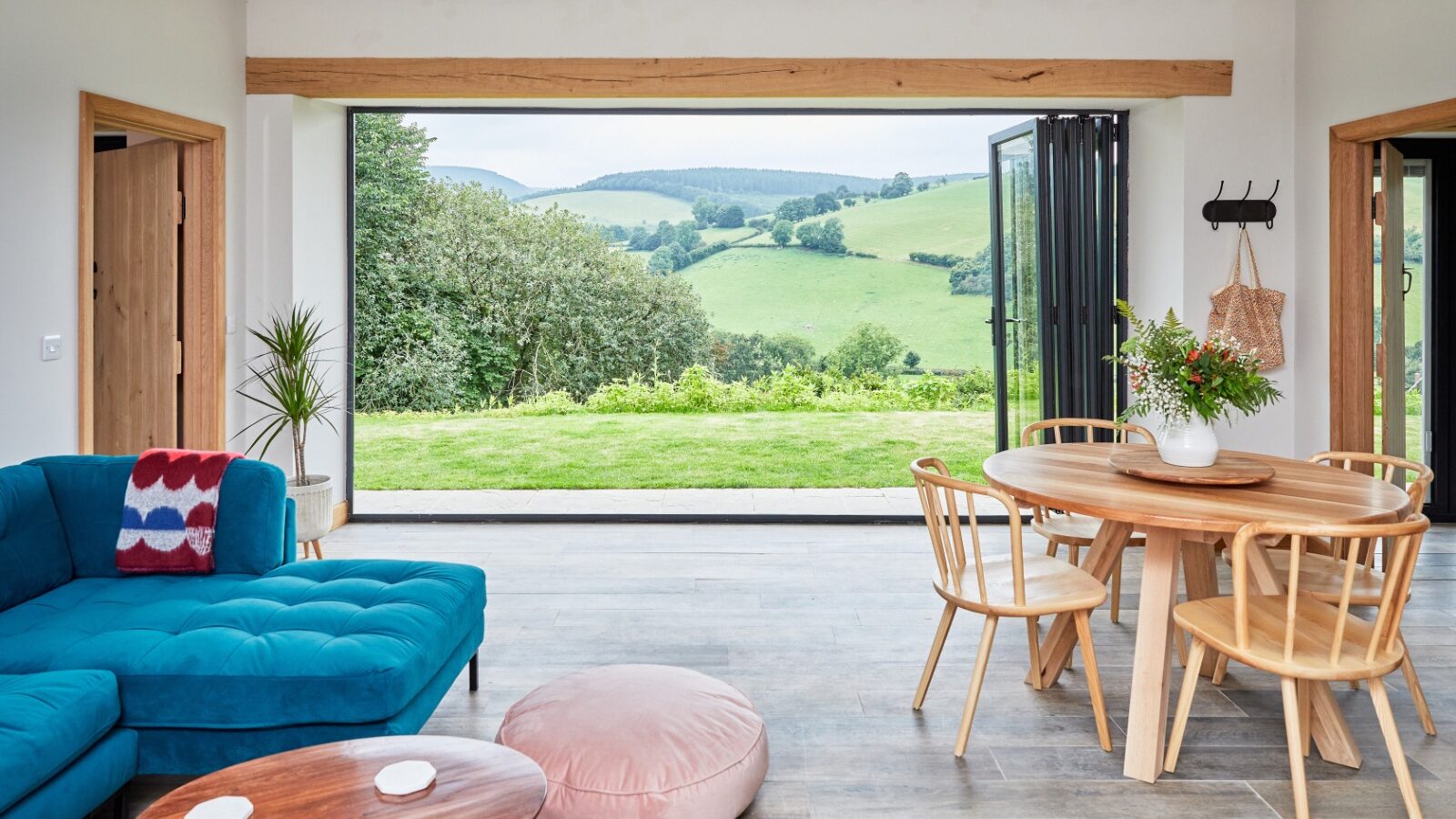 A modern living room with a Burrows blue sofa, round wooden table, and chairs. Large bi-fold doors open to a scenic view of green hills. A pink floor cushion and Dolassey plants decorate the space, enhancing the bright and airy atmosphere.