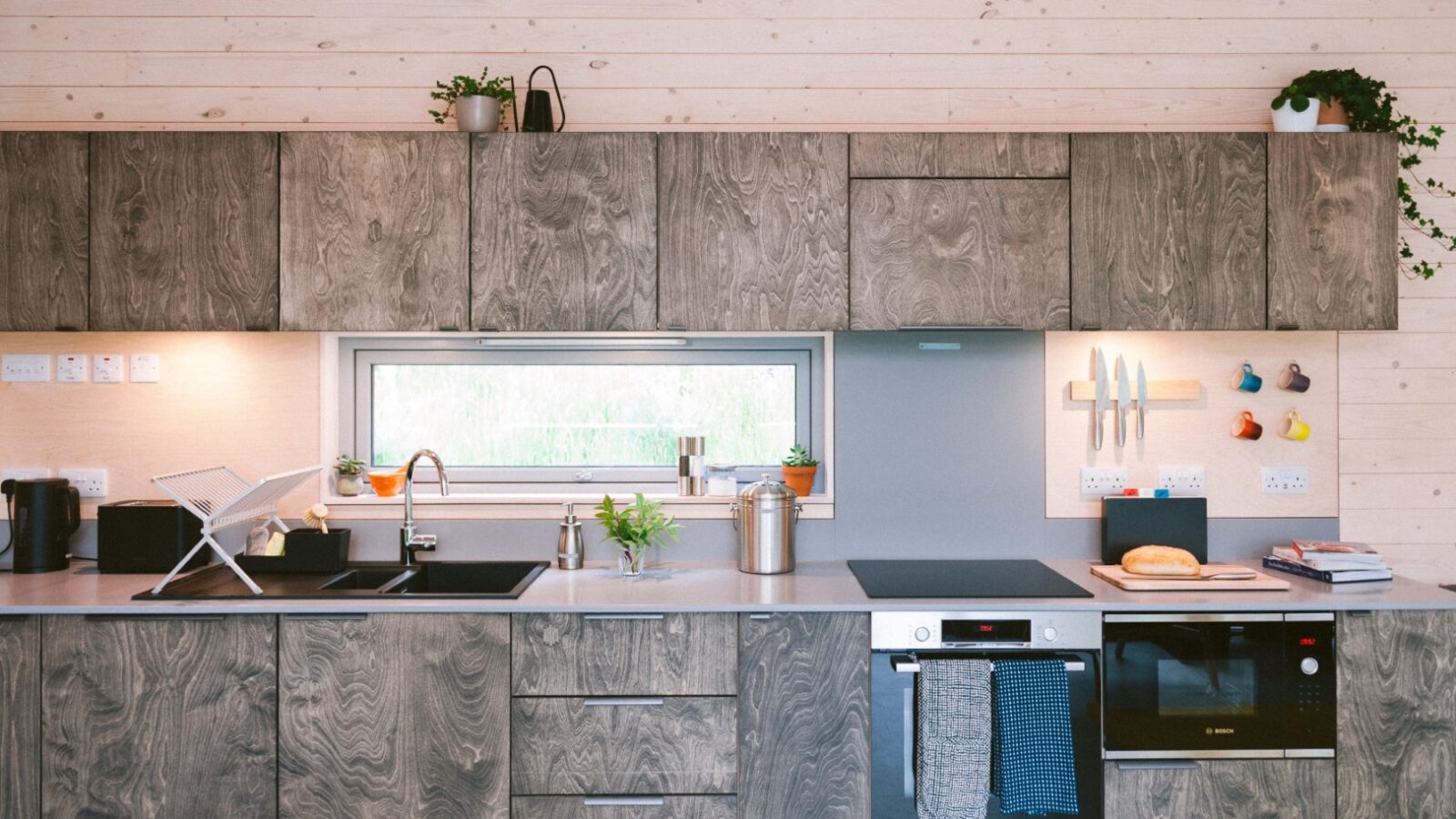 A modern kitchen with wooden cabinets reminiscent of cozy cabins features a built-in oven, stove, and a wall-mounted knife holder. Plants adorn the space above the cabinets. A small window above the sink provides natural light, while various kitchen items are neatly arranged on the counter.