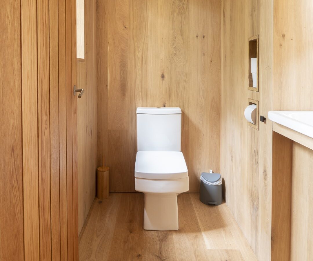 A modern bathroom features a toilet centered against a wooden wall with a wooden floor, reminiscent of a cozy treehouse. To the right of the toilet is a roll of toilet paper in a built-in holder and a small gray trashcan on the floor. To the left is a wooden door. Natural light enters from windows.