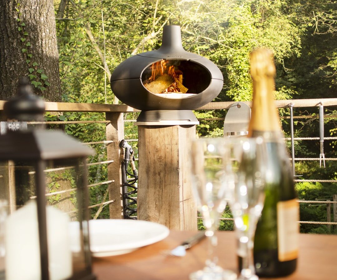 A cozy outdoor setting with a wood-fired oven burning on the deck of a charming treehouse. In the foreground, a wooden table is set with a lantern, glasses, a plate, utensils, and a bottle of champagne. The background features lush green trees bathed in sunlight.