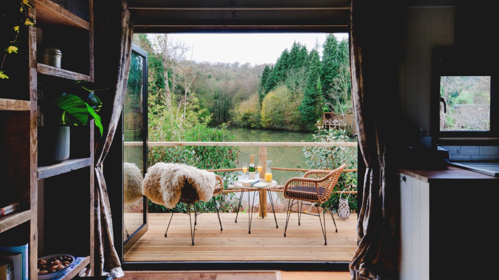 A cozy shepherds hut interior opens to a wooden deck with two chairs and a table overlooking a serene lake surrounded by trees, creating the perfect retreat.