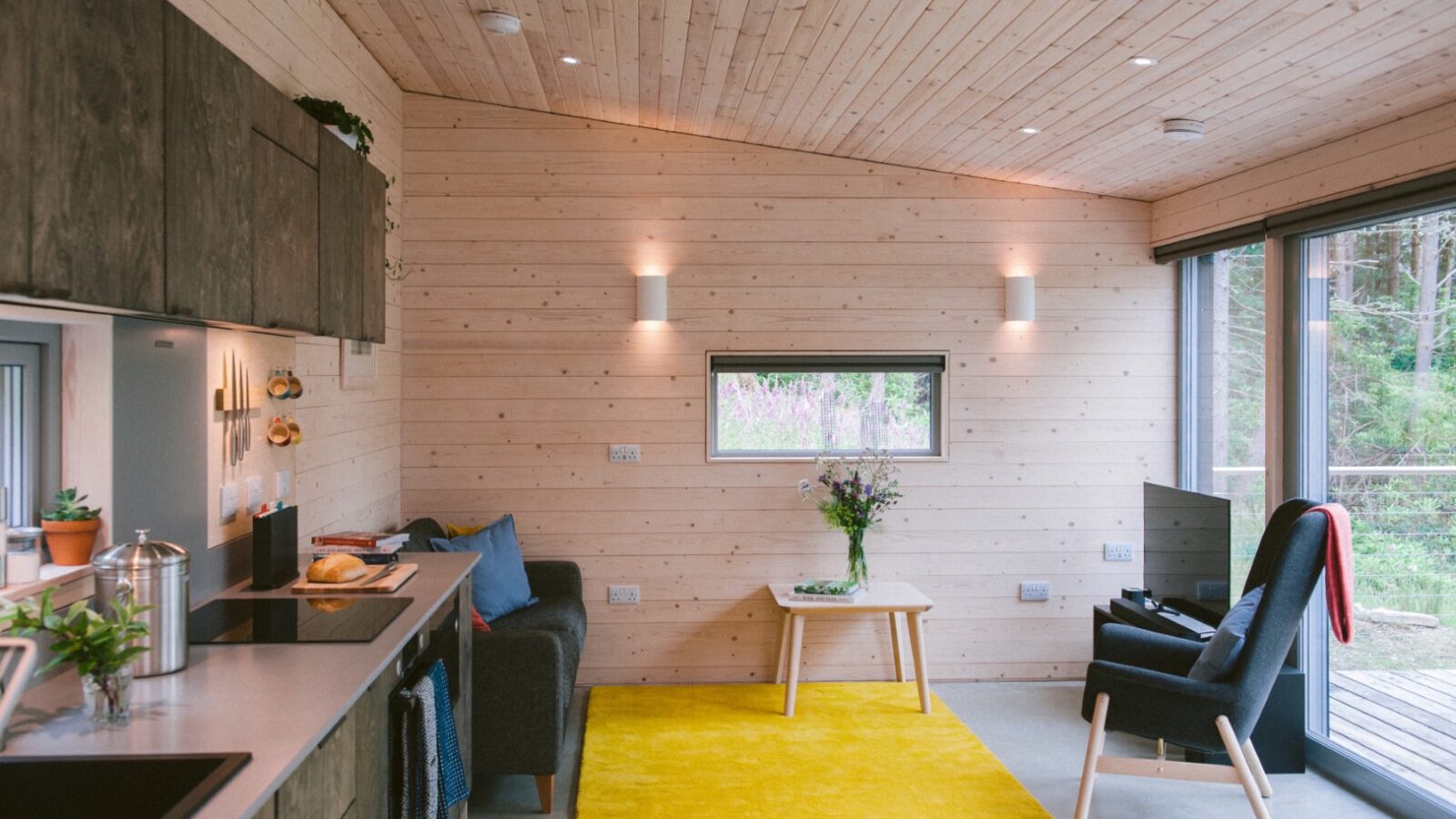 A cozy, modern living area in an ecotone cabin with wooden walls and ceiling, featuring a small kitchen, a black armchair, a table with flowers, and a bright yellow rug. Large glass doors open to a deck with a breathtaking forest view.