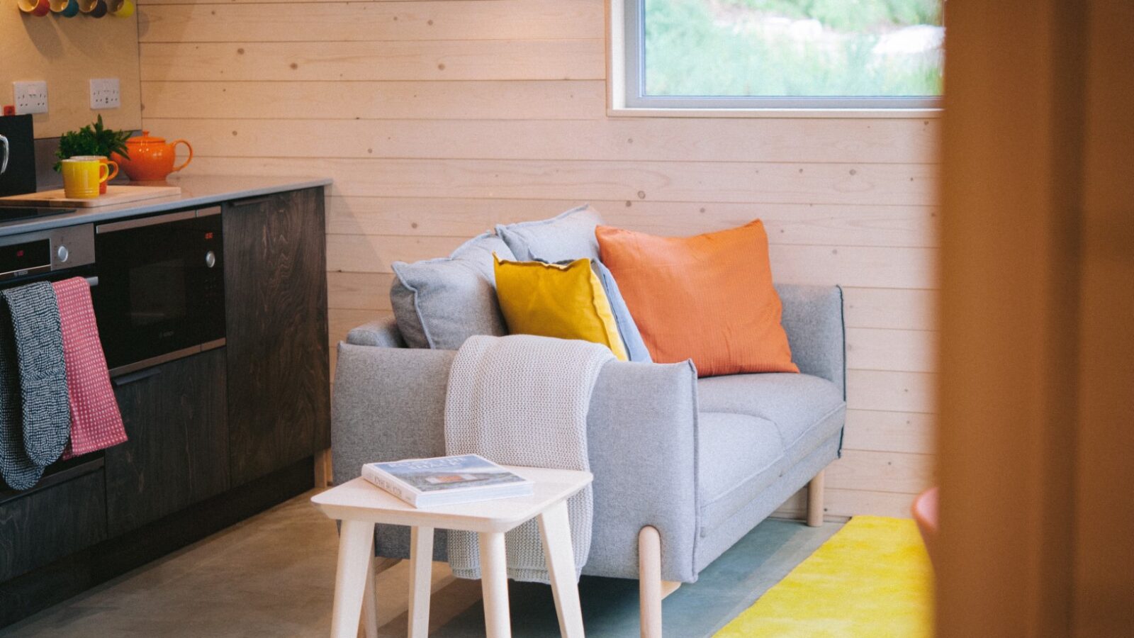 A cozy cabin living space features a gray sofa adorned with orange and yellow pillows, a light wooden side table with a book, and a small kitchen area with colorful mugs hanging above the stove. The ecotone of light wood-paneled walls complements the bright window perfectly.