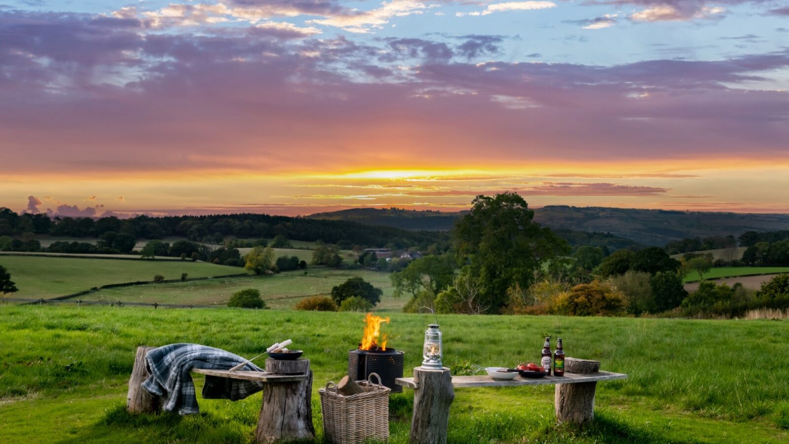 Experience the enchanting sunset view at Moonrise Lodges, nestled in a countryside landscape with a cozy fire pit, wooden benches, and a charming picnic setup on the grass.