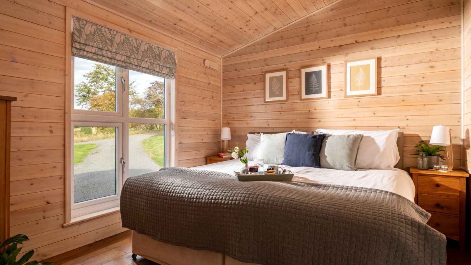 Experience the tranquility of Moonrise Lodges in a cozy wooden bedroom. Enjoy a large bed adorned with decorative pillows and bedside tables, perfect for unwinding. A tray with drinks awaits by the window, offering a serene nature view that invites relaxation.