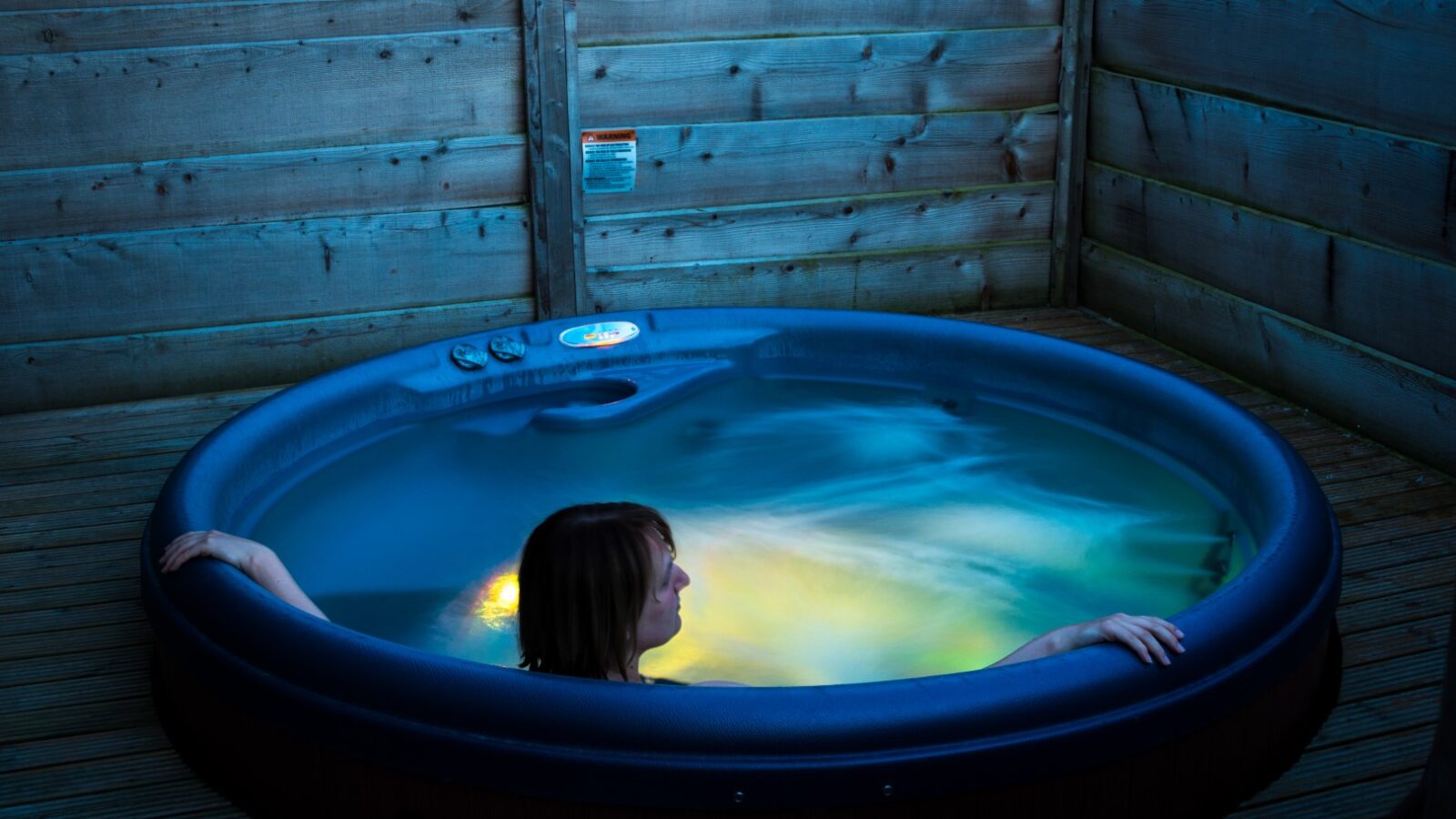 A person unwinds in a circular hot tub, embraced by wooden walls and bathed in colorful water lighting. As the moonrise casts its glow, the serene atmosphere feels like an escape to tranquil lodges.