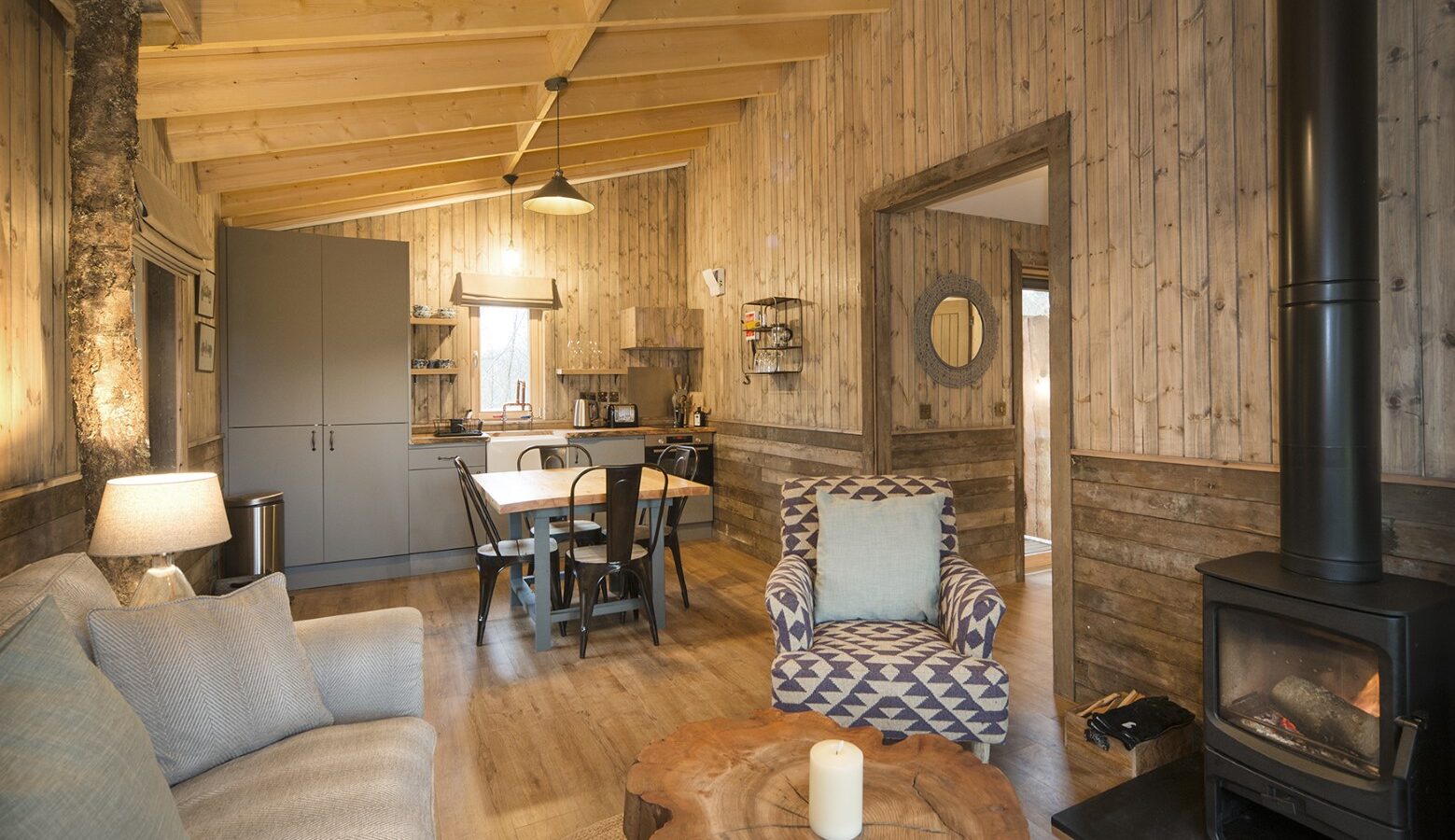 A cozy treehouse interior features a living area with a sofa, an armchair, and a wood-burning stove. The dining area and kitchen are visible, with wooden walls and a beamed ceiling creating a warm, rustic atmosphere—a perfect treehouse rental retreat.