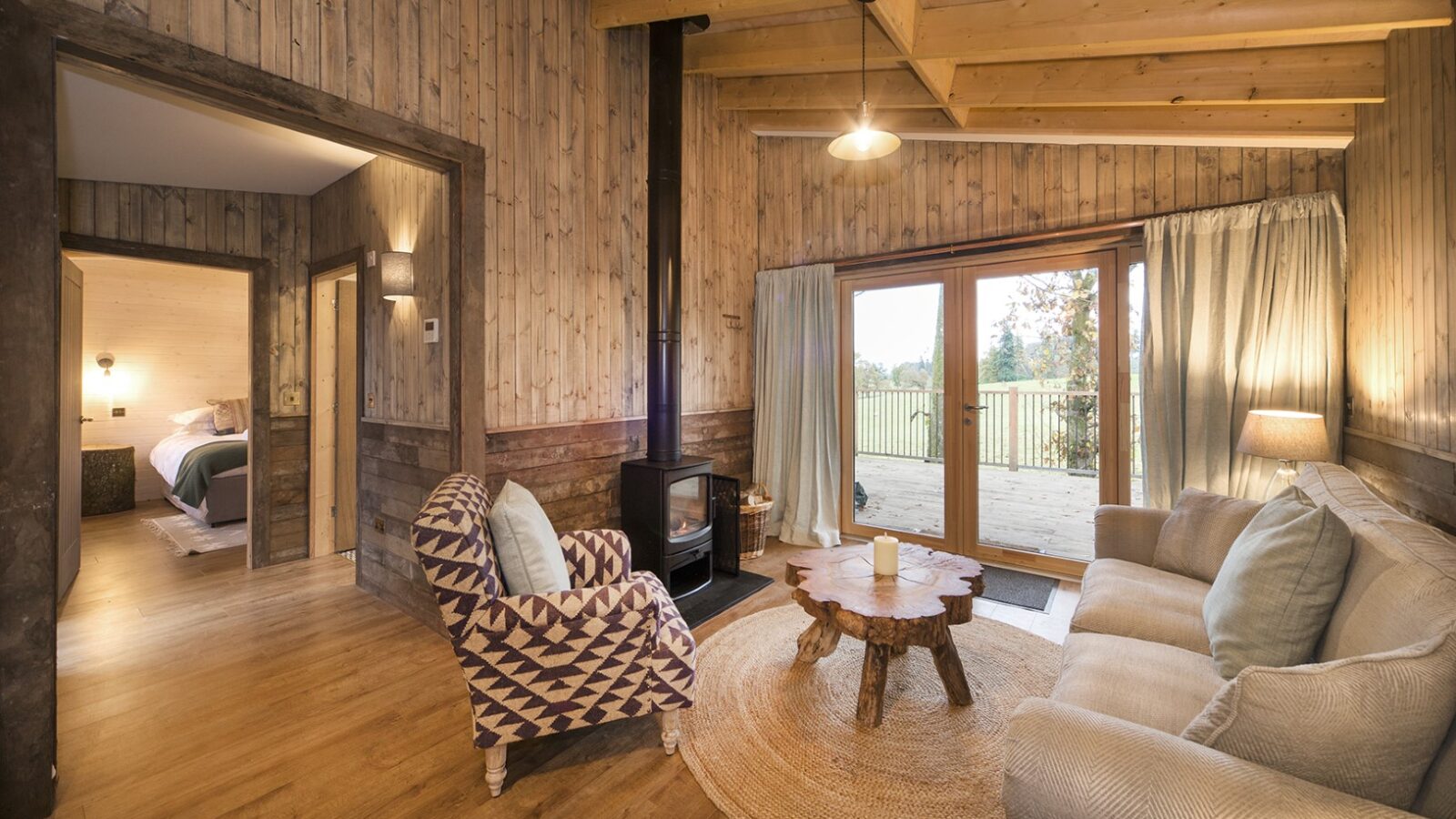 A cozy cabin interior with wooden walls and ceiling feels like a charming treehouse. A sofa faces a wood-burning stove and a rustic coffee table, while a patterned armchair sits nearby. Large windows and a door lead to a deck with views of trees, enhancing the treehouse rental vibe.