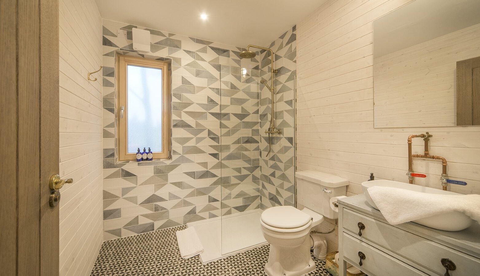 The modern bathroom evokes the charm of a treehouse, with geometric tile design. A shower with a glass partition stands on the left, beside a frosted window. On the right are a toilet and vintage-style sink with mirror, all grounded by black and white patterned tiles.