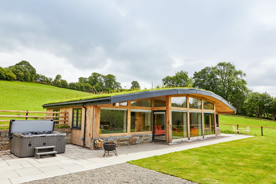 A modern, single-story house with a green roof and large glass windows sits nestled in the lush, rural landscape of Burrows. A hot tub graces the patio, and grassy hills and trees stretch into the background under a cloudy sky.