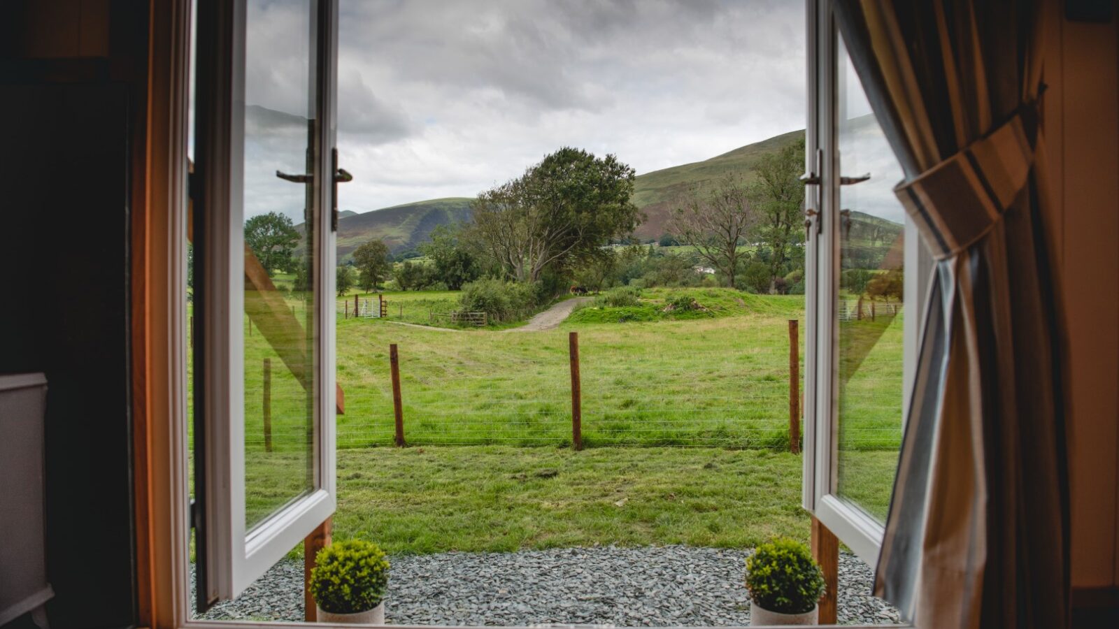 Open double doors reveal a view of The Vale, where a grassy field with rolling hills and a cloudy sky stretches into the distance, all elegantly framed by curtains and small shrubs.