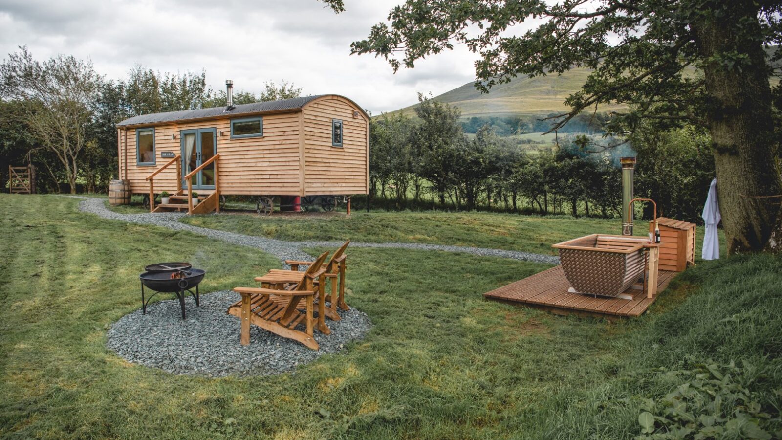 Wooden cabin nestled in the grassy landscape of The Vale, featuring a fire pit, two chairs, and a small hot tub under a tree with hills painting the background.