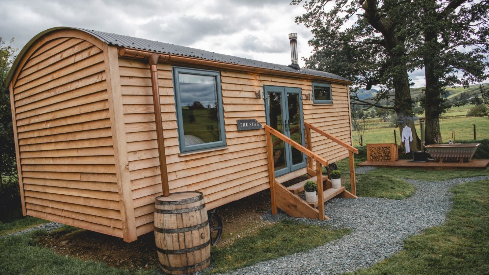 A charming wooden cabin with a curved roof and stairs nestled in the lush greenery of The Vale, surrounded by a tree, a barrel, and a picturesque landscape beyond.