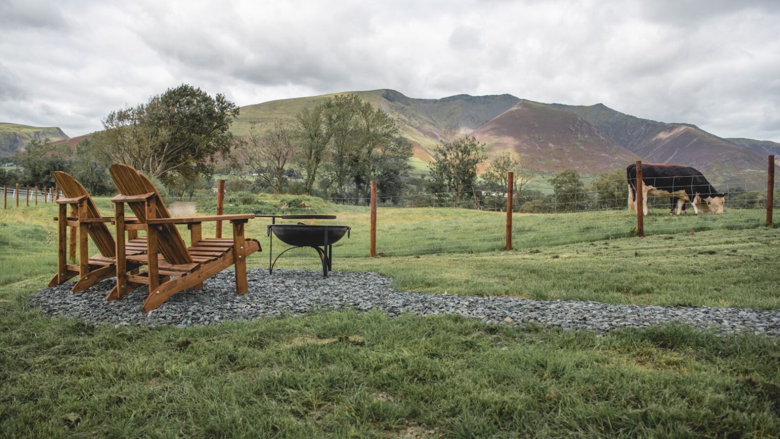 Two wooden chairs face a fire pit on a gravel path, overlooking The Vale where cows graze in a lush green field with mountains in the background.