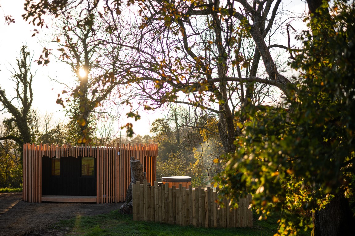 A charming wooden cabin, nestled among trees, basks in the warm glow of a setting sun. A small fenced area with a round hot tub enhances the scene, while autumn leaves and a serene pond view add color to this peaceful retreat.