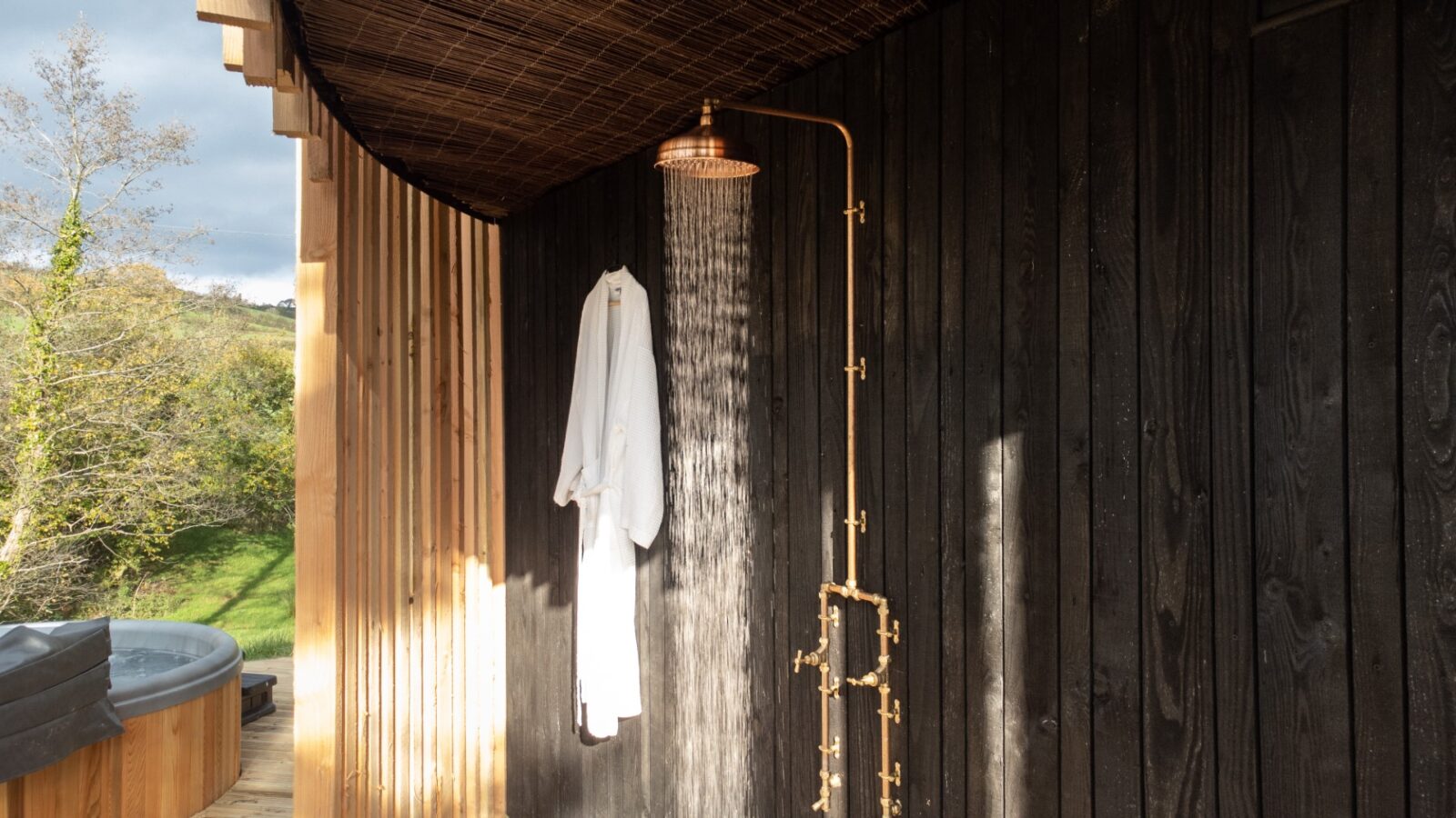 Outdoor shower area at Pond View Lodges features a copper shower head and dark wood paneling. A white robe hangs nearby, while in the background, lush greenery and a hot tub complete the serene setting.