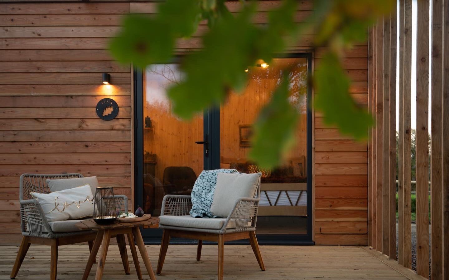 A cozy outdoor patio with two wicker chairs, each adorned with cushions and a throw blanket. A small wooden table between the chairs holds a tray, creating the perfect spot to enjoy a serene pond view. Warm light shines from a wall-mounted lamp, while a tree branch partially frames the scene.