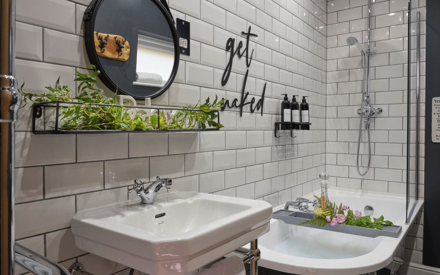 A stylish bathroom in the cosy Cowshed features white subway tiles, a round mirror, and a shelf adorned with greenery. A bathtub filled with water, flowers, and a book nestles under the rain shower. Black 