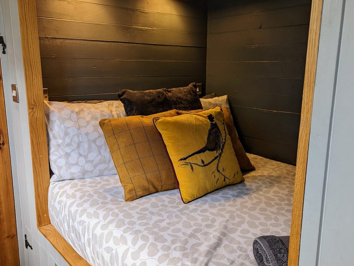 Cozy bedroom nook with a small bed framed by wooden panels, reminisciscent of the quaint charm found in Shepherds huts. The bed is adorned with white and grey patterned sheets, a black cushion, and yellow pillows featuring bird designs. Two wall-mounted lamps provide warm lighting. A small animal picture hangs above.