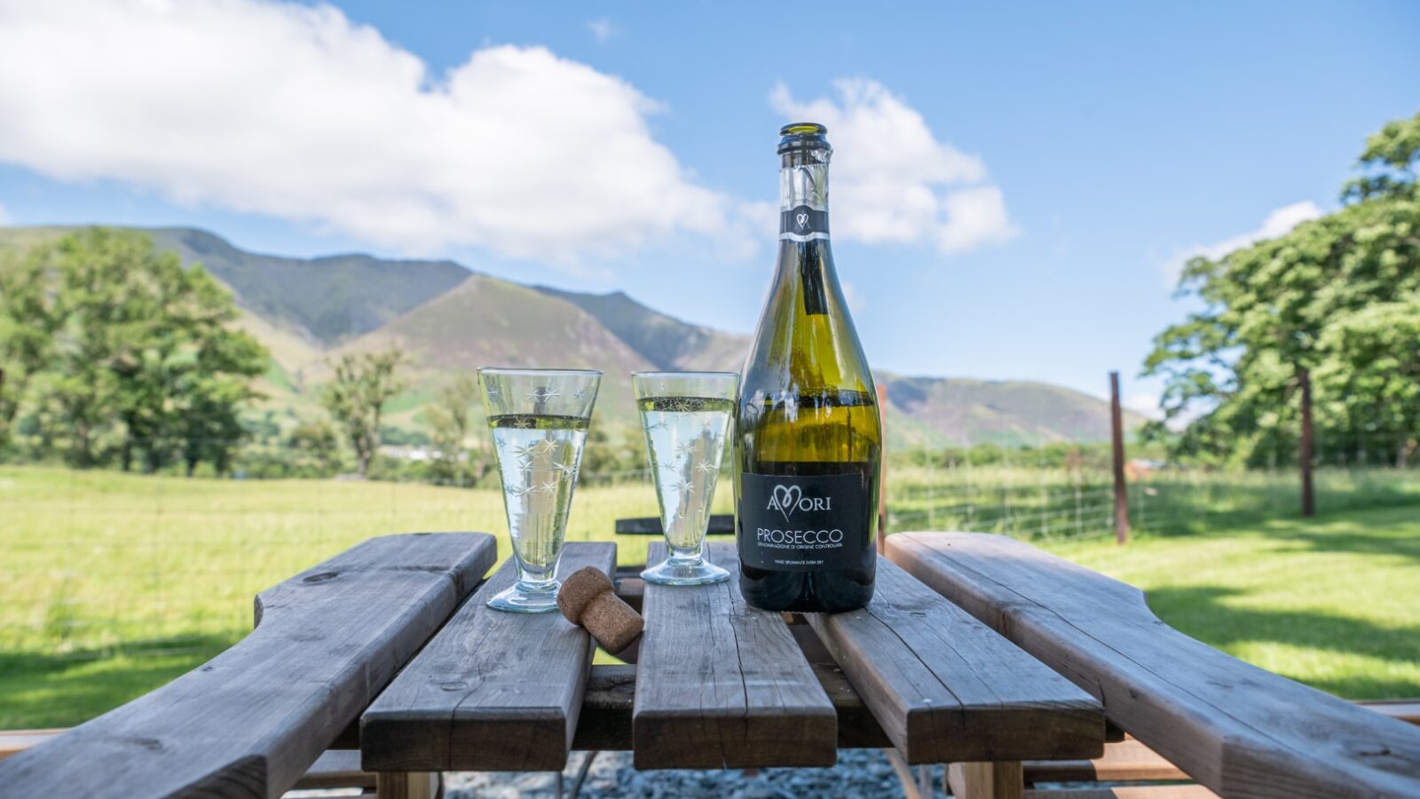 A bottle of prosecco and two filled glasses sit on a wooden table outdoors, with the majestic mountains and lush grassy field of The Vale in the background.